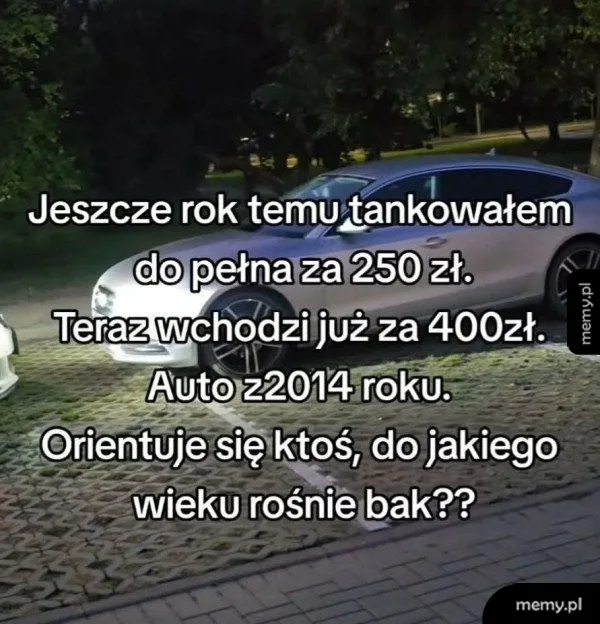 Srebrny samochód osobowy nocą na parkingu z tekstem o cenach paliwa.